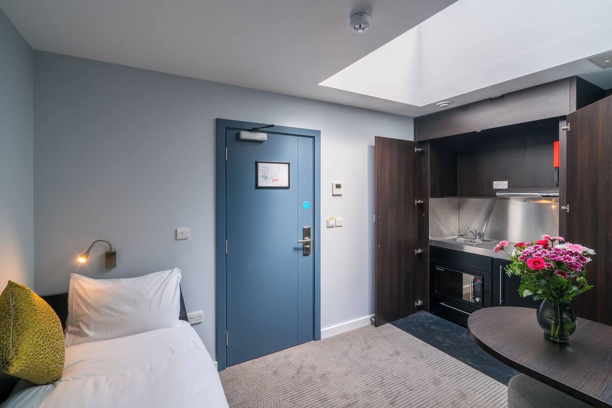 Ett modernt hotellrum med en säng, ett skrivbord, en blå dörr, ett litet kök och ett takfönster.