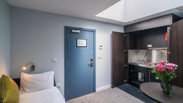 Et moderne hotelværelse med seng, skrivebord, en blå dør, tekøkken og ovenlys.