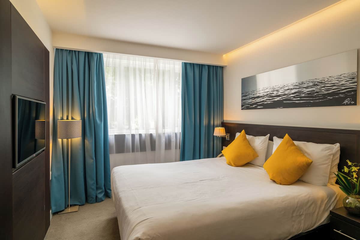 Ett hotellrum med en stor säng, två gula kuddar, en tavla som föreställer ett havsskepp och en tv på vänster sida.