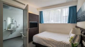Ett hotellrum med en säng, ett nattduksbord med en lampa, ett fönster med gardiner, en tv och ett badrum med handfat och spegel.
