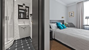 A modern bathroom with a glass shower enclosure, a white sink, and a mirror. The room has a tiled floor and a large bed with a grey headboard and blue pillows. - 15th Avenue Hotel (Vilnius)