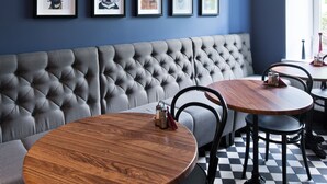 A room with a blue wall, framed dog portraits, a tufted bench, and round wooden tables. - 15th Avenue Hotel (Vilnius)
