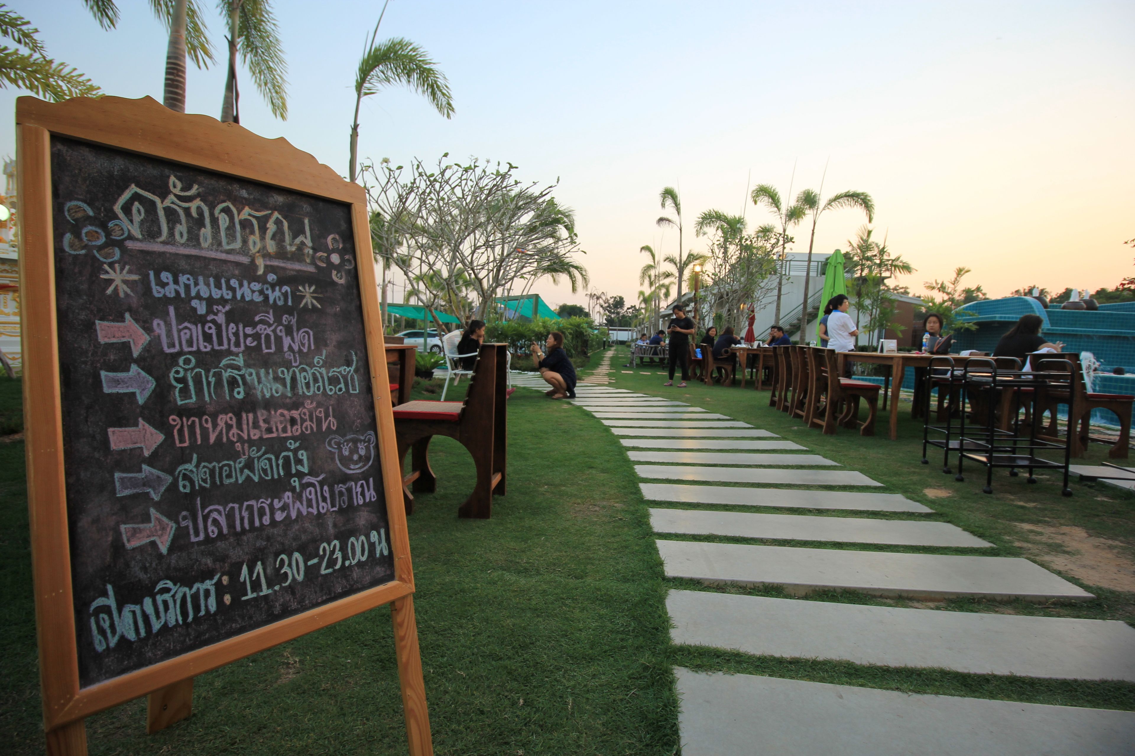 An outdoor seating area with tables and chairs, a chalkboard with text, and palm trees in the background.