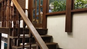 Una escalera de madera con varios peldaños y barandillas, que conduce a una zona de descanso con vistas a plantas de interior y una persona al fondo.