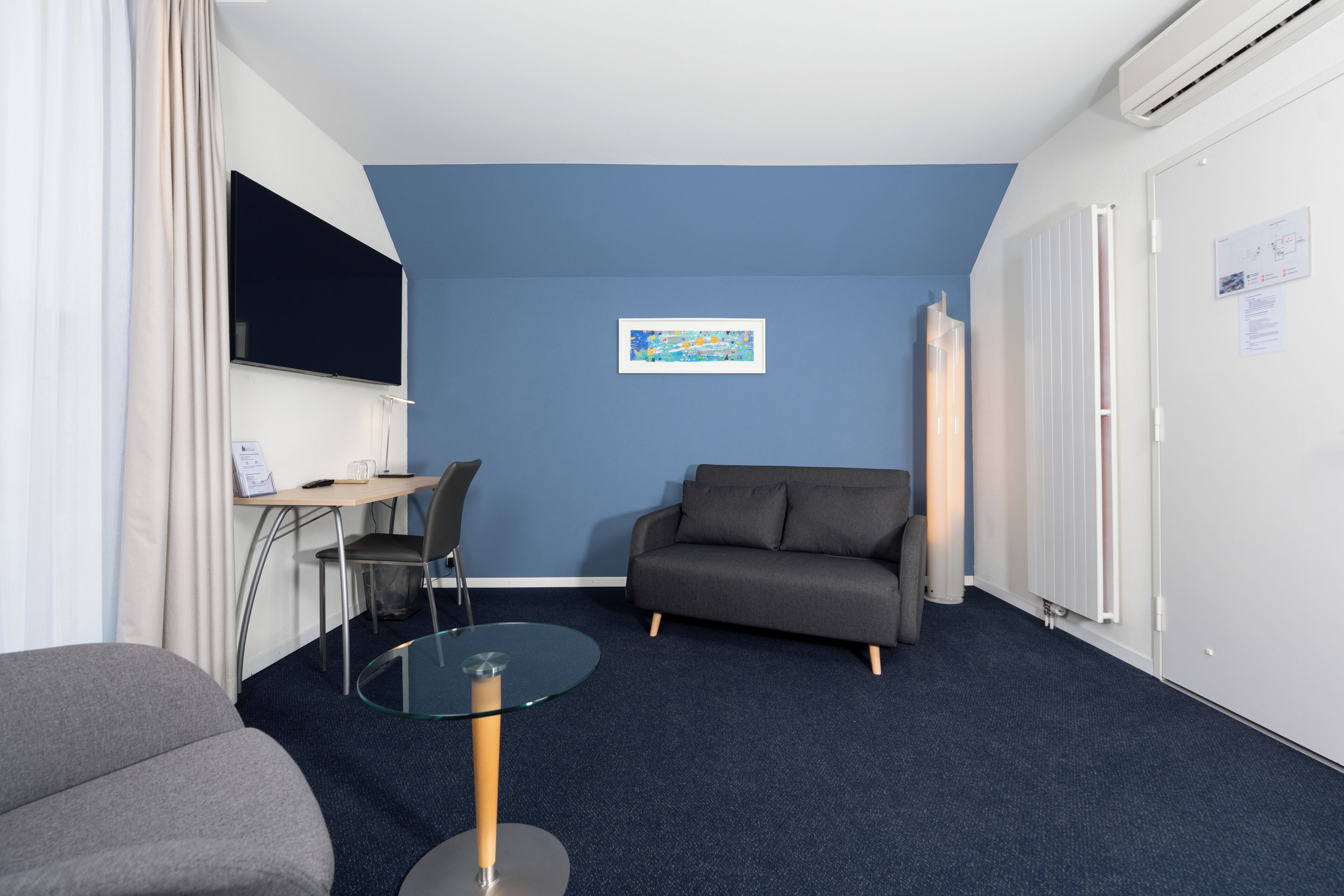 Une pièce moderne avec un mur d’accent bleu, un canapé gris, une table en verre, un téléviseur et un radiateur.