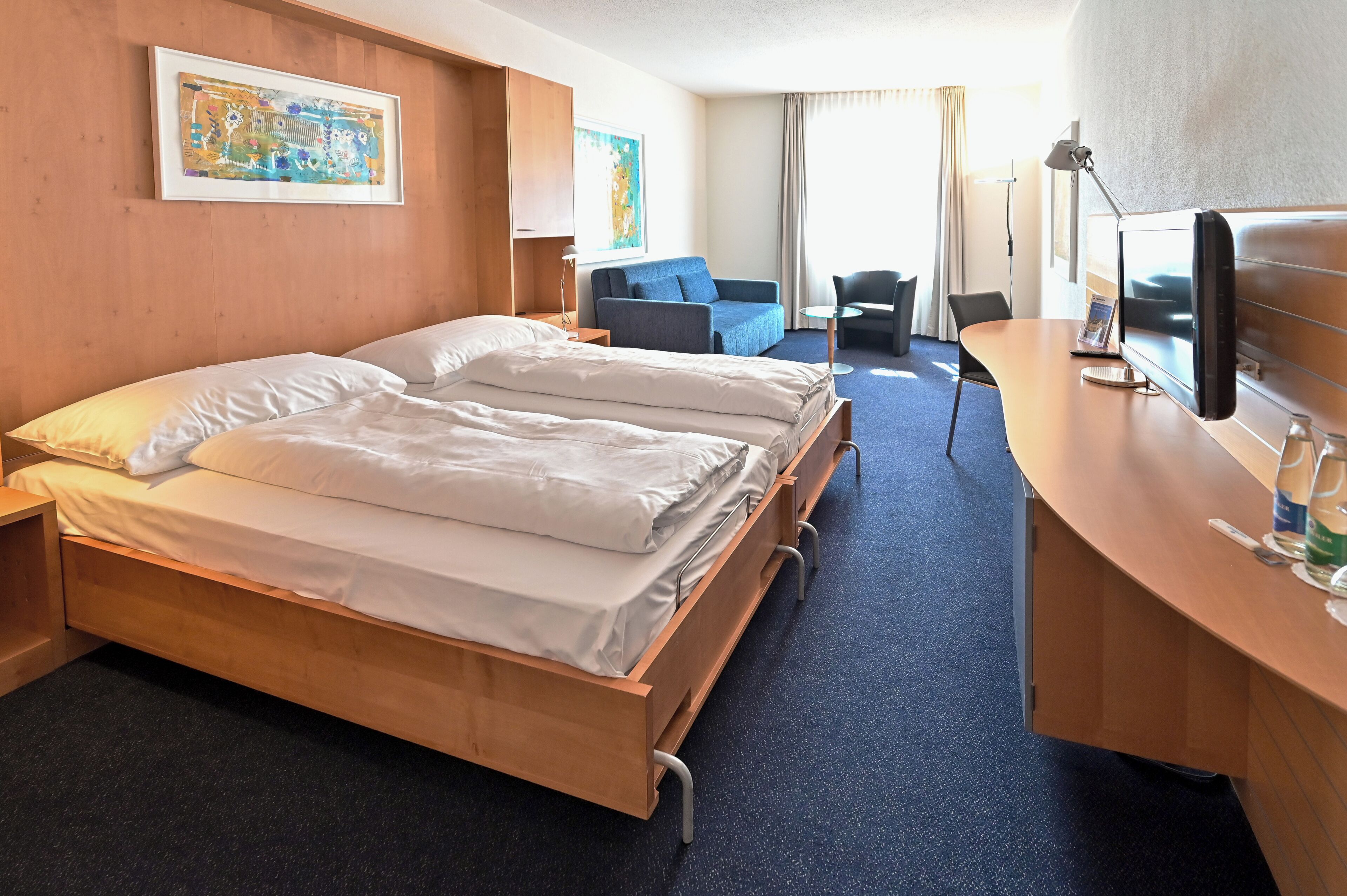 A hotel room with a bed, a desk, a chair, a TV, and a painting on the wall.
