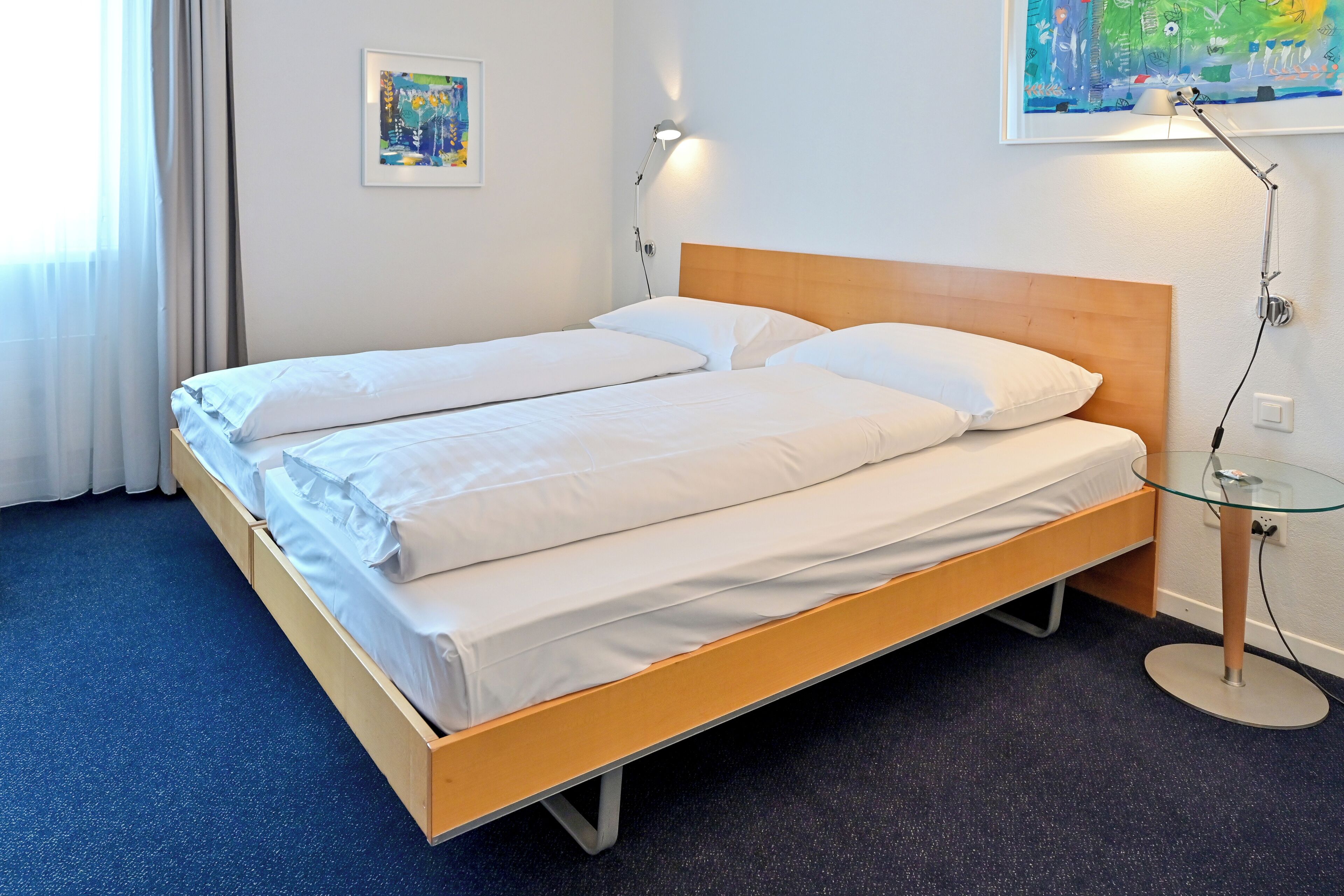 Cama doble con sábanas blancas, cabecera de madera, mesita de noche con lámpara y un cuadro en la pared.