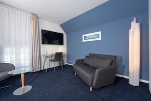 Una habitación de hotel moderna con una pared azul de acento, un sofá gris, una mesa de centro de vidrio, un escritorio con una silla y un televisor de pantalla plana montado en la pared.