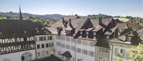 Un edificio europeo tradicional con una torre destacada, un patio con un pequeño edificio, y vista a una colina boscosa.