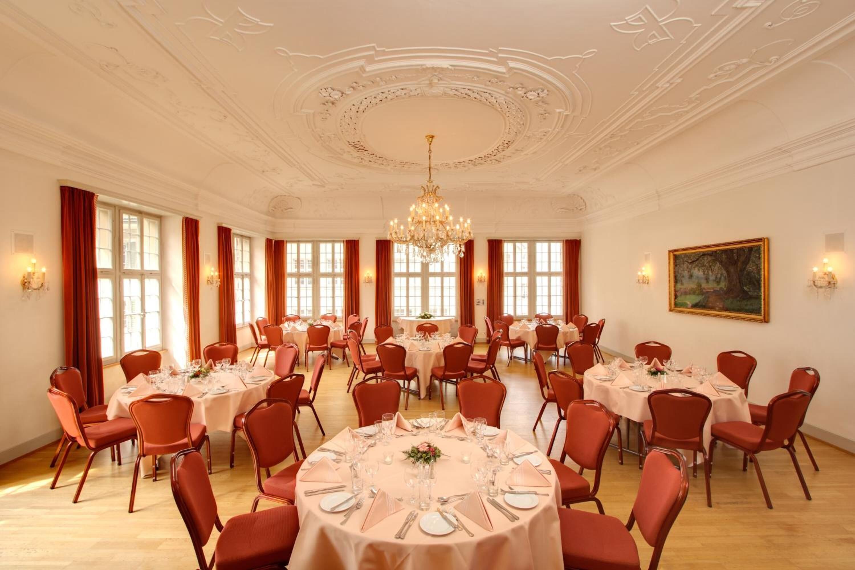 Un amplio salón de banquetes con mesas redondas dispuestas para el servicio de comidas, sillas rojas, candelabros y grandes ventanales.