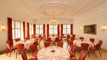 Un amplio salón de banquetes con mesas redondas dispuestas para el servicio de comidas, sillas rojas, candelabros y grandes ventanales.
