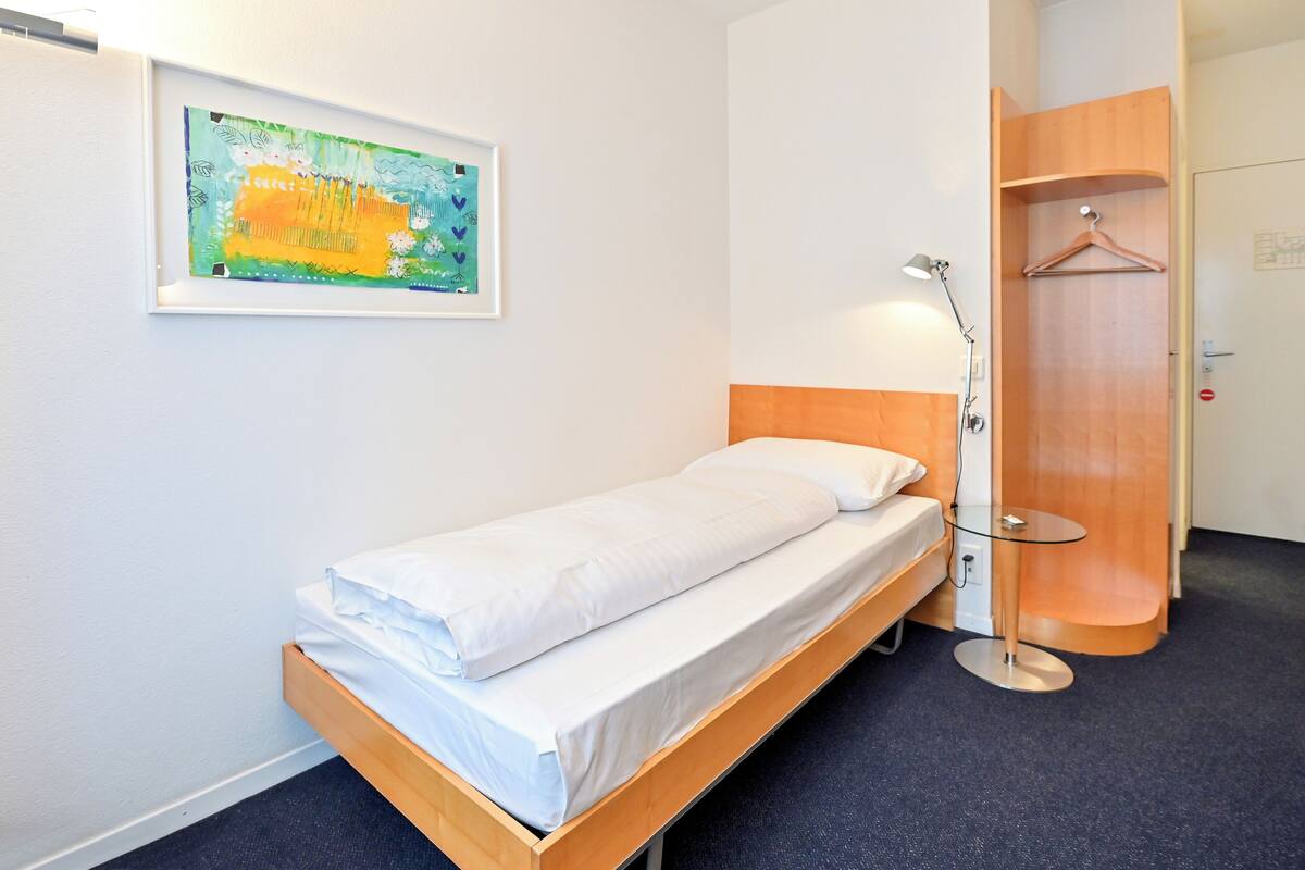 A single bed with white linens, a bedside table with a lamp, a wooden wardrobe, and a framed abstract painting on the wall.
