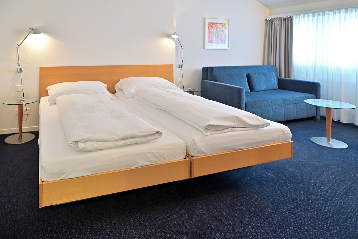 A hotel room with a double bed, a blue armchair, a small table, and two bedside lamps.