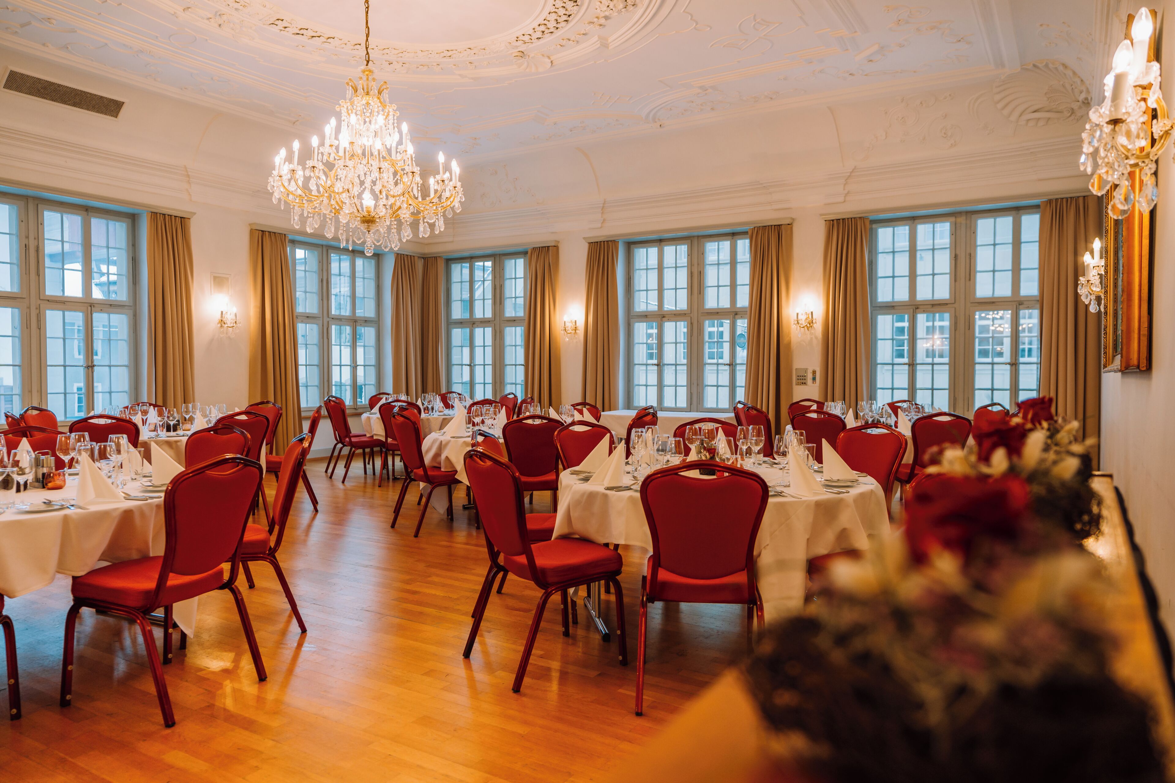 A banquet hall with multiple tables set for dining, red chairs, and a chandelier.