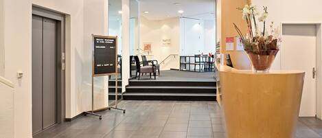 A modern hotel lobby with a reception desk, a staircase leading to the upper floor, and a sign indicating the location.
