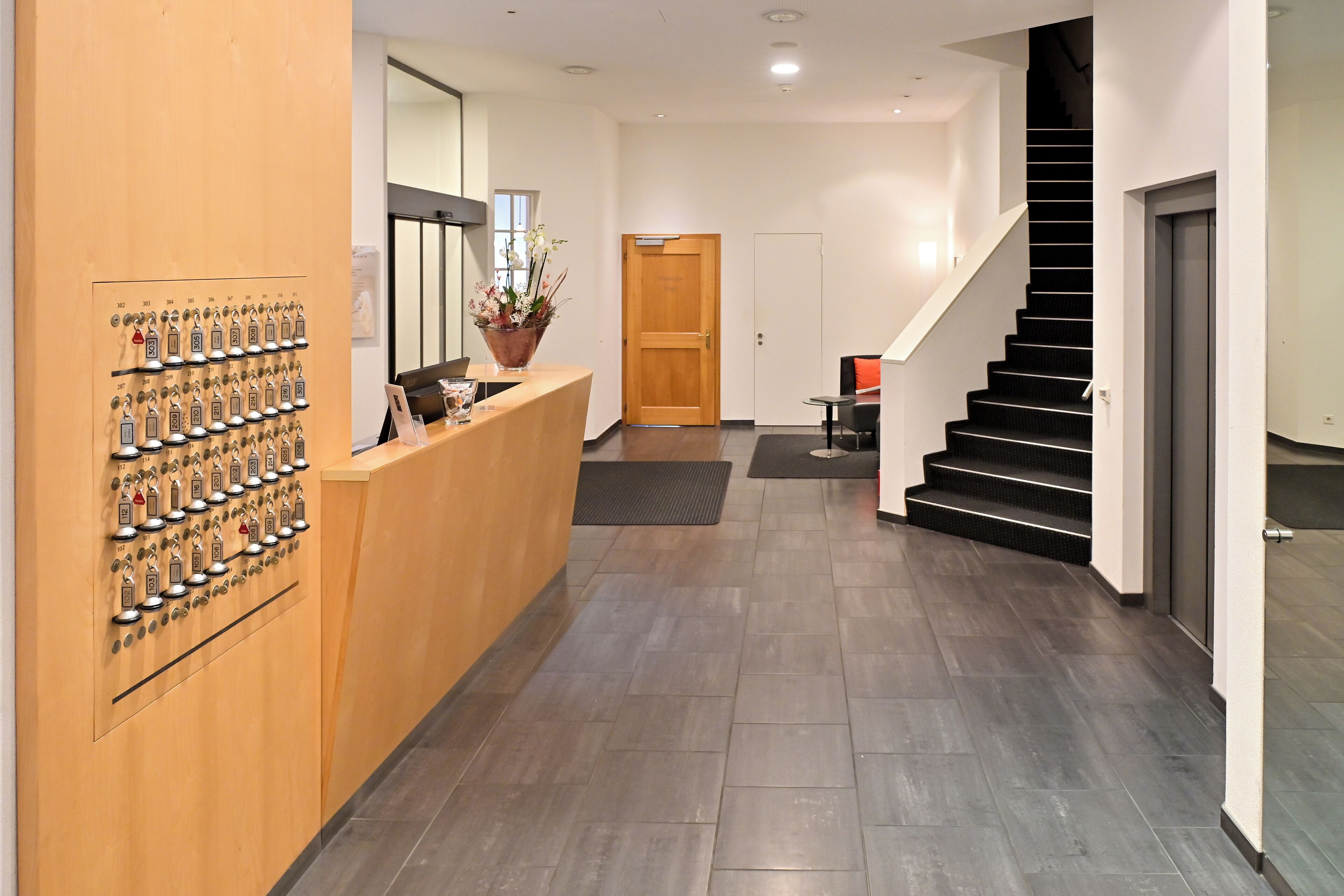 A hotel reception area with a wooden reception desk, a wall-mounted keycard system, a staircase, and a door leading to another room.