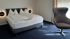 A neatly made bed with white linens and a dark headboard, a modern armchair, and a wall-mounted light fixture. - Hotel Zofingen (Zofingen)