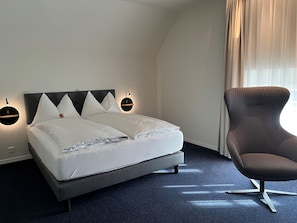A neatly made bed with white linens and a dark headboard, a modern armchair, and a wall-mounted light fixture. - Hotel Zofingen (Zofingen)