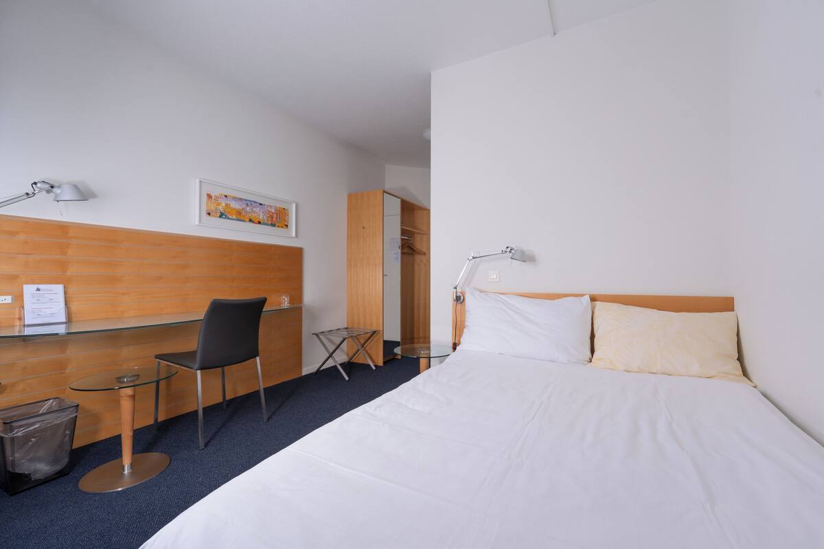 A hotel room with a bed, a desk, a chair, a small table, a trash bin, and a wardrobe.
