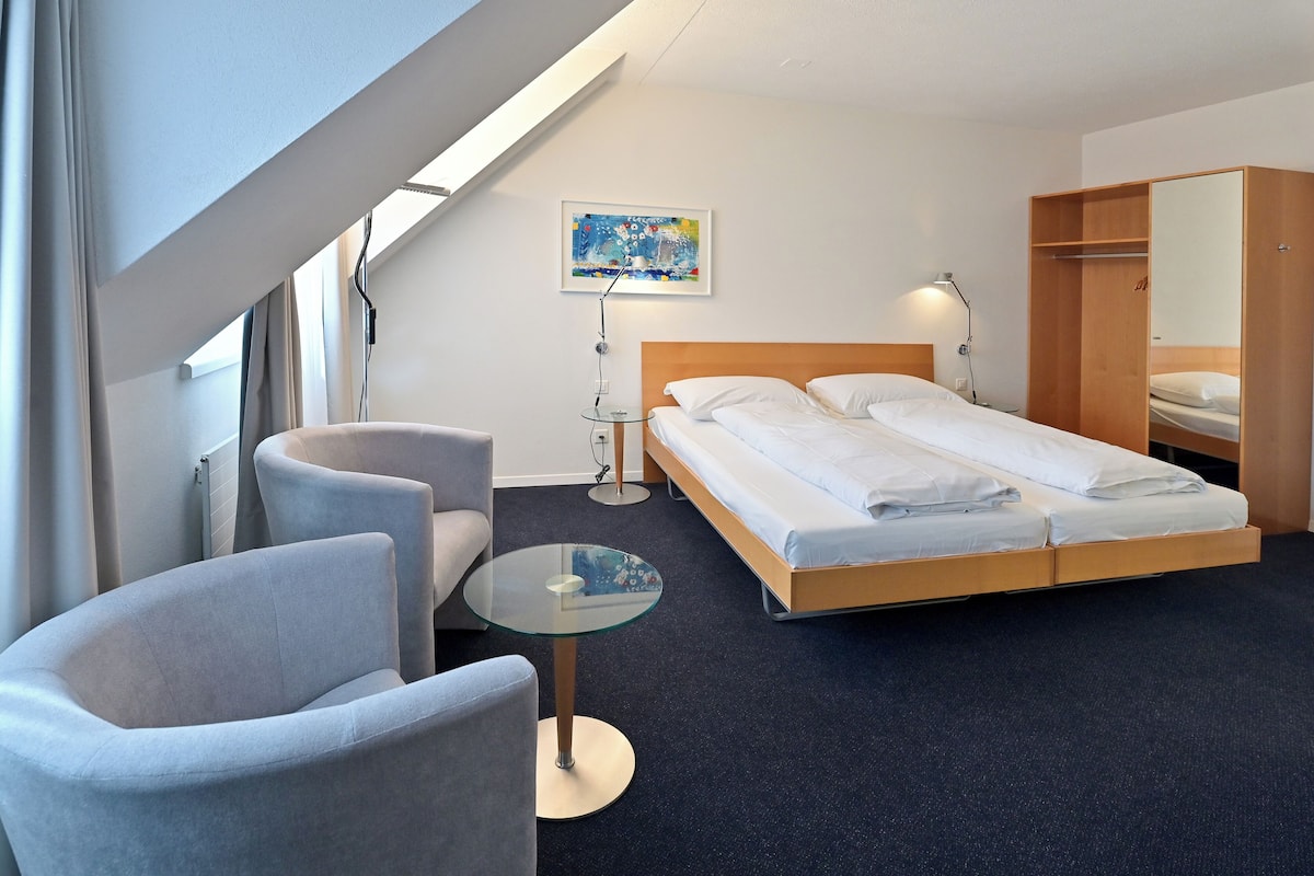 A modern hotel room with a bed, two armchairs, a small round table, a ceiling light, and a painting on the wall.