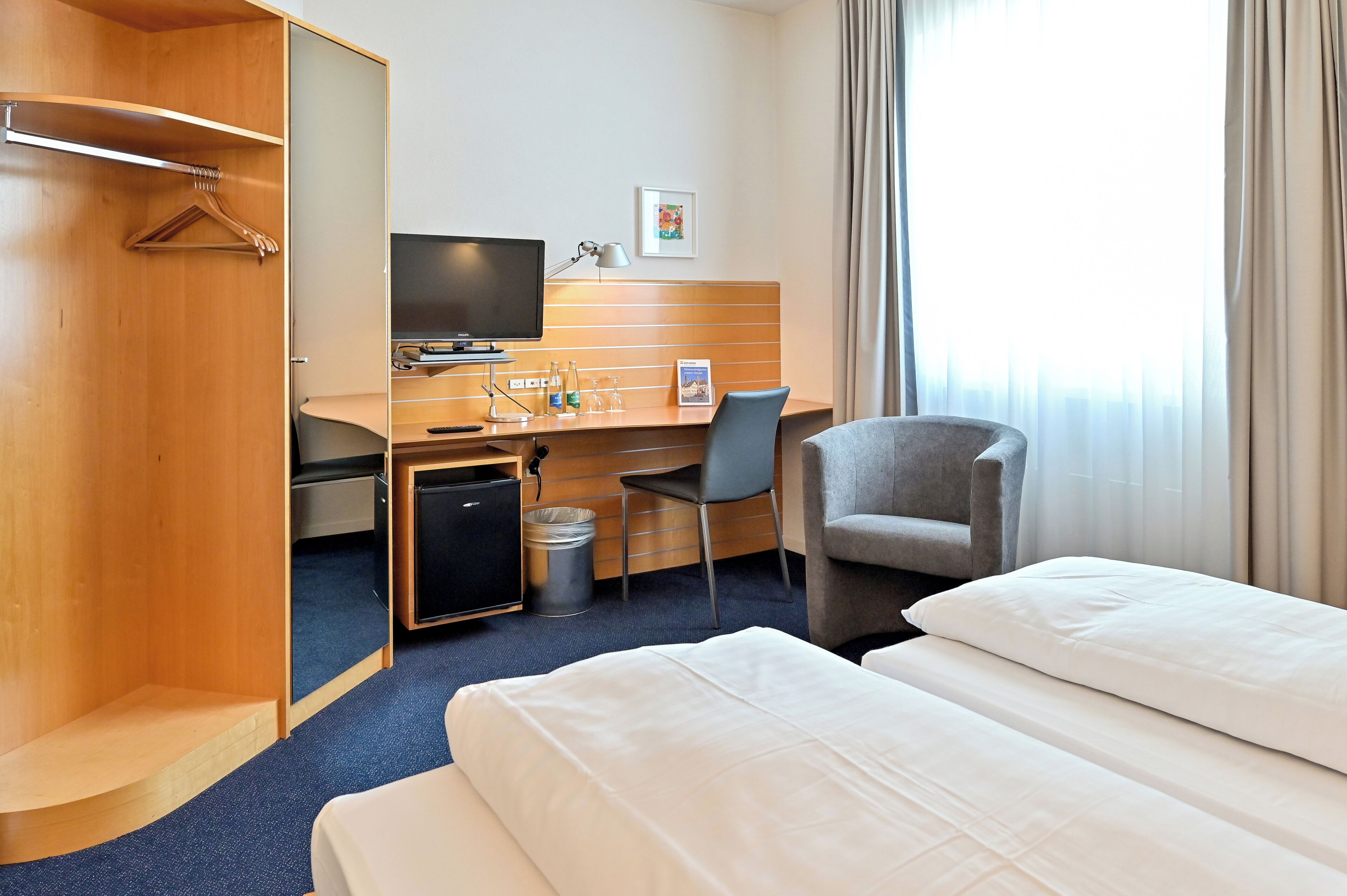 Habitación de hotel con cama, escritorio con computadora, silla, armario y ventana con cortinas.