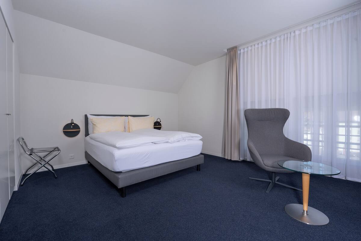 A hotel room with a bed, a chair, a glass table, and curtains.