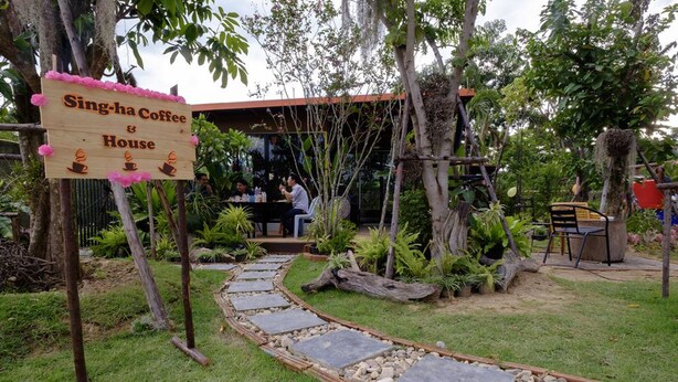 Un letrero de madera anuncia Singha Coffee & House, un jardín con un camino de piedra y mesas al aire libre.