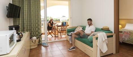 A small, cozy room with a green sofa, a wooden cabinet with a microwave, and a television mounted on the wall.