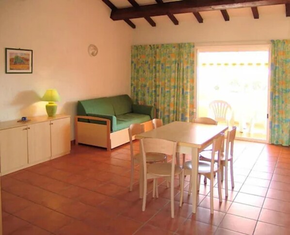 Una sala de estar con un sofá verde, una mesa de comedor con sillas, una lámpara y una ventana con cortinas.