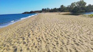 A wide sandy beach with gentle waves, a clear sky, and a few buildings in the background.