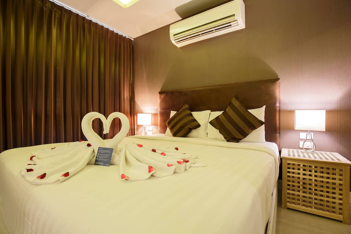 Una cama cuidadosamente tendida con toallas con forma de corazón y pétalos de rosa, una mesita de noche de madera con una lámpara y un aire acondicionado montado en la pared.