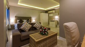 A modern living room with a sofa, a wooden coffee table with a floral centerpiece, a chair, and a small kitchenette area with a refrigerator and a microwave.