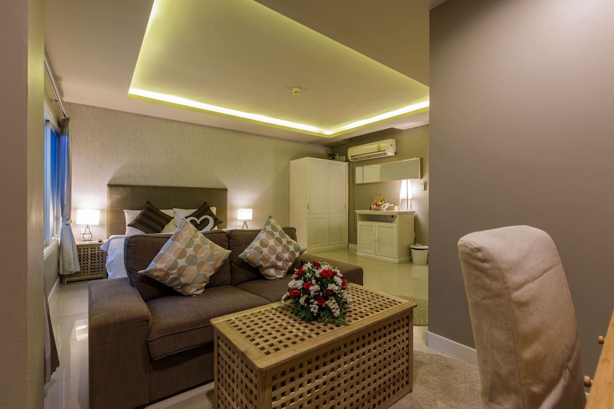 Una sala de estar moderna con un sofá, una mesa de centro de madera con un centro de mesa floral, una silla y una pequeña zona de kitchenette con refrigerador y microondas.