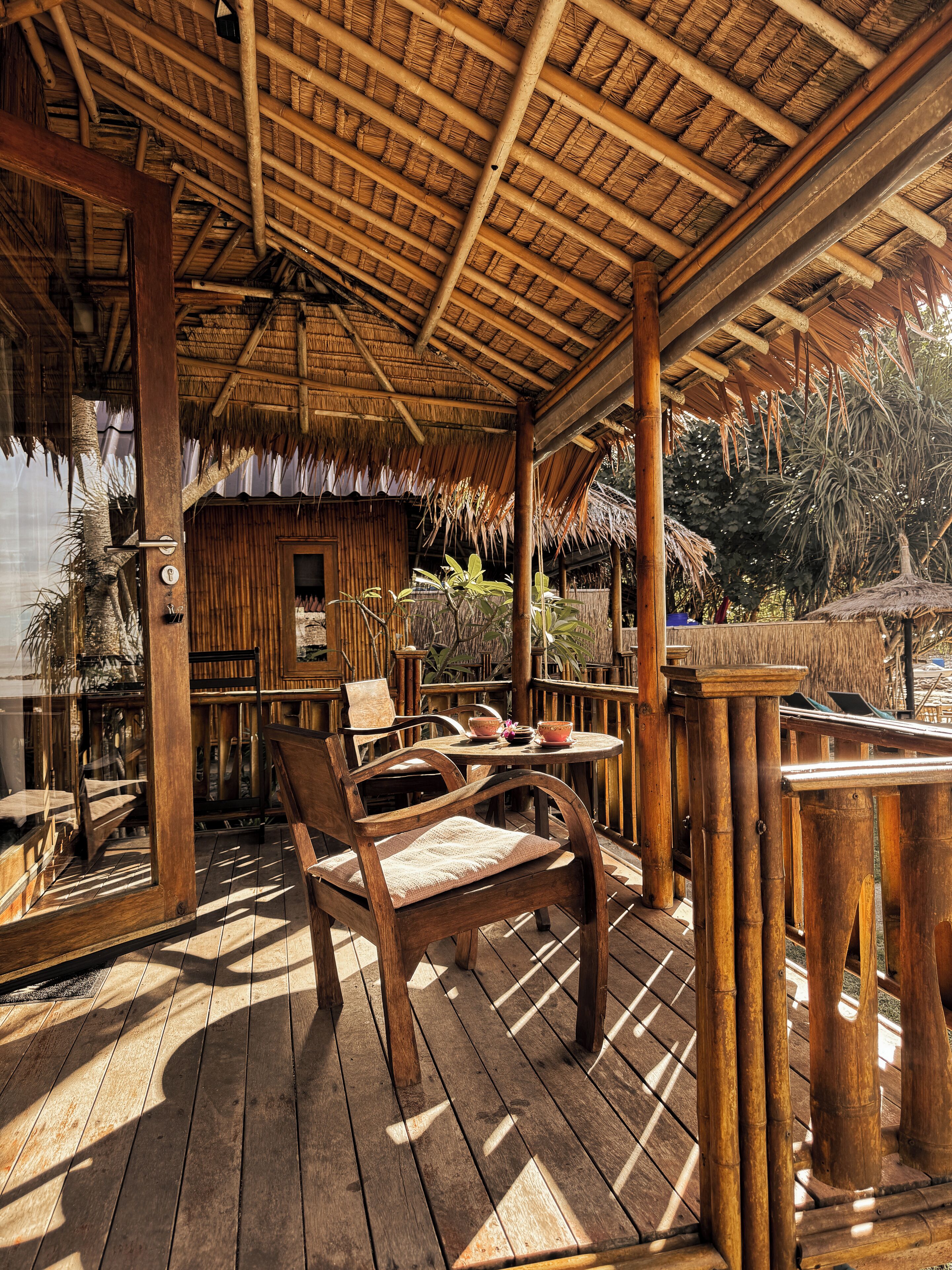 Una terraza de madera con techo de paja, sillas de madera y una mesa con cuencos y una vela.