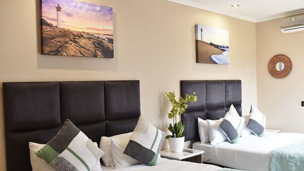 A hotel room with two beds, a large painting of a lighthouse, a potted plant, and a round mirror on the wall. - Regal Inn Gateway Umhlanga (Umhlanga)