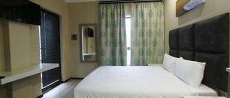A hotel room with a bed, a bedside table, a TV mounted on the wall, and a window with curtains.