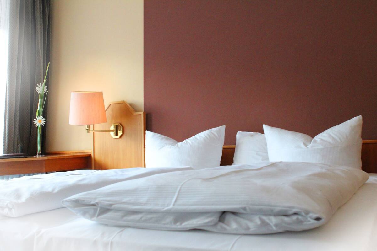 Cama bem arrumada com lençóis e almofadas brancas, cabeceira de madeira e luminária fixada na parede.