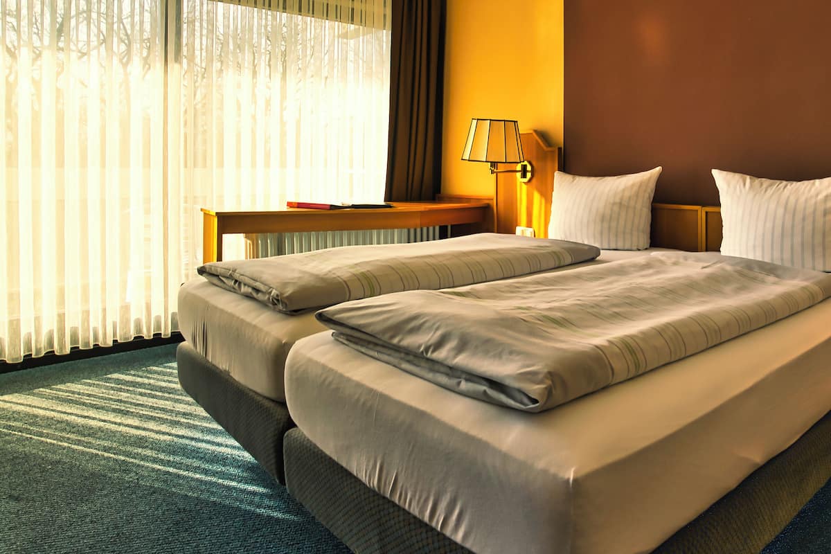 Quarto de hotel com uma cama grande, mesa de cabeceira, abajur e vista para o exterior através de cortinas translúcidas.
