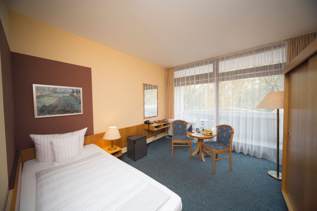 Quarto de hotel com cama, escrivaninha, cadeira, abajur e janela com cortinas.