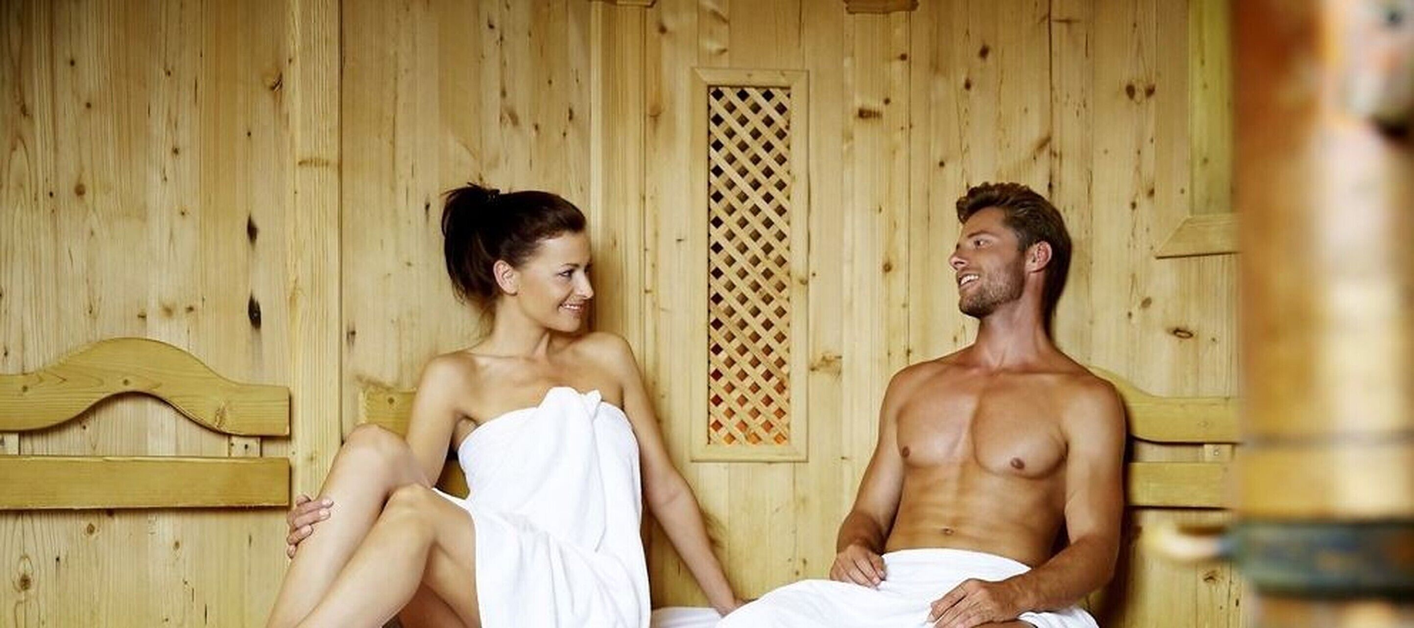 A wooden sauna room with two benches, one occupied by a person and the other by a person with a towel wrapped around their body.