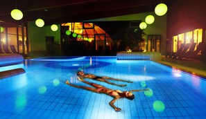 A swimming pool with two people floating on their backs, surrounded by lounge chairs and a building with large windows in the background. -  Reichels Parkhotel (Bad Windsheim)