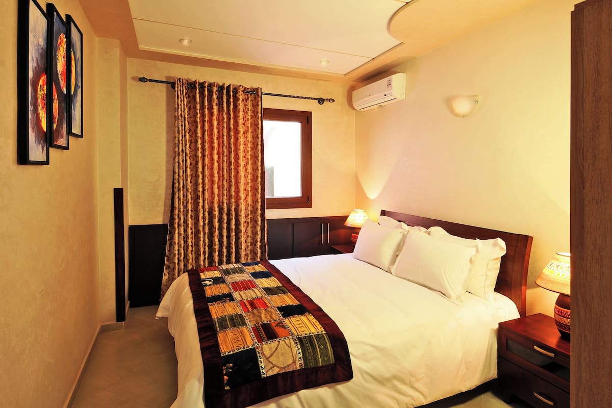 Una habitación de hotel con cama, mesita de noche, lámpara, aire acondicionado y ventana con cortinas.