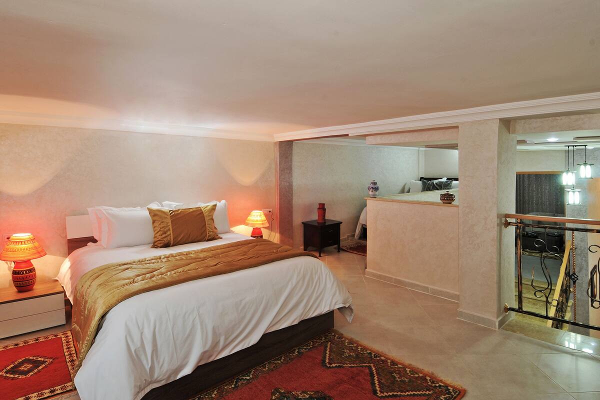 Habitación de hotel con cama, mesitas de noche, alfombra roja y baño visible a través de una puerta abierta.