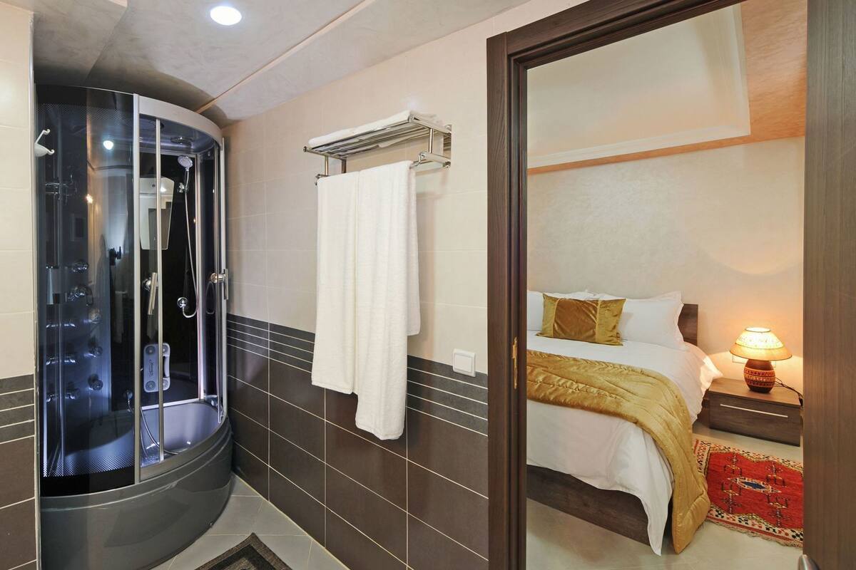 Un baño moderno con ducha envuelta en vidrio, un perchero y una cama con mesita de noche.