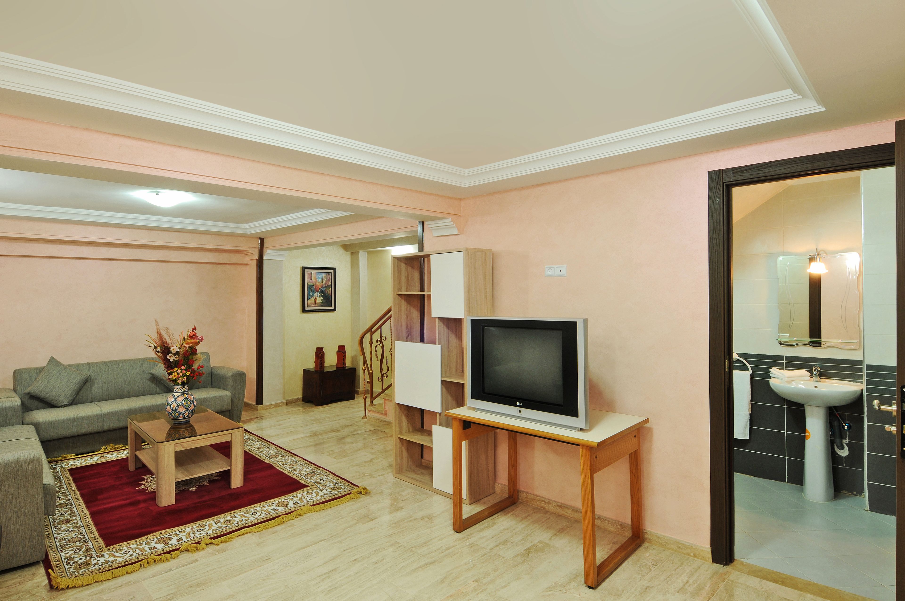 Una sala de estar con sofá, una mesa de centro con un jarrón, televisor sobre un mueble bajo y una escalera que lleva a otra habitación.