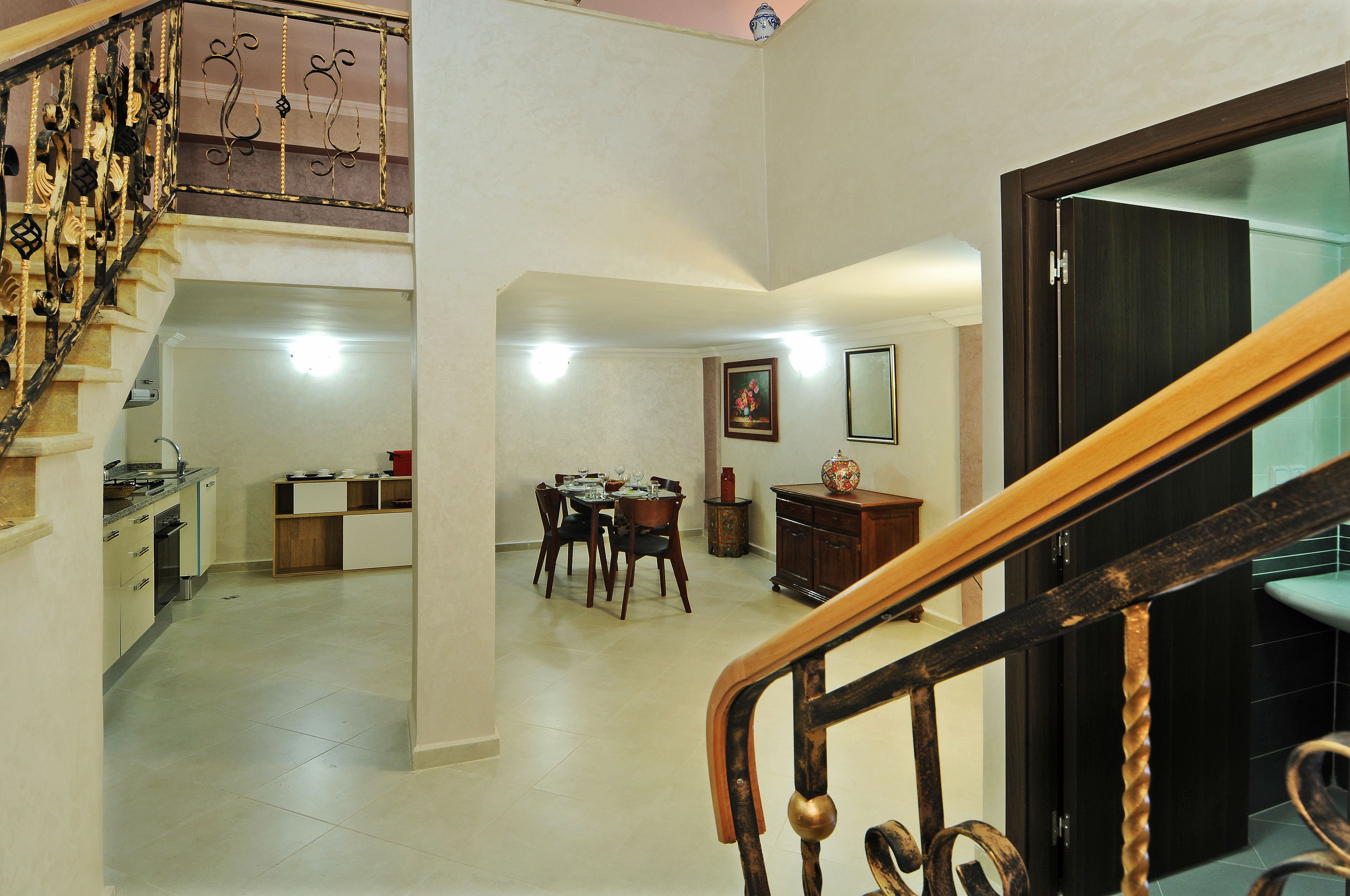 A spacious living area with a dining table and chairs, a staircase with ornate railings, and a kitchen in the background.