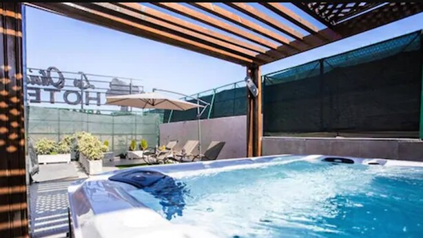 Un jacuzzi con una pérgola de madera encima, un área de descanso con sillones y sombrillas, y vista a un edificio con un letrero.