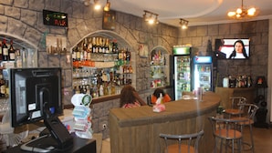 A bar with a stone wall, shelves of bottles, a TV screen, and bar stools. - Hotel Saphir (Abidjan)