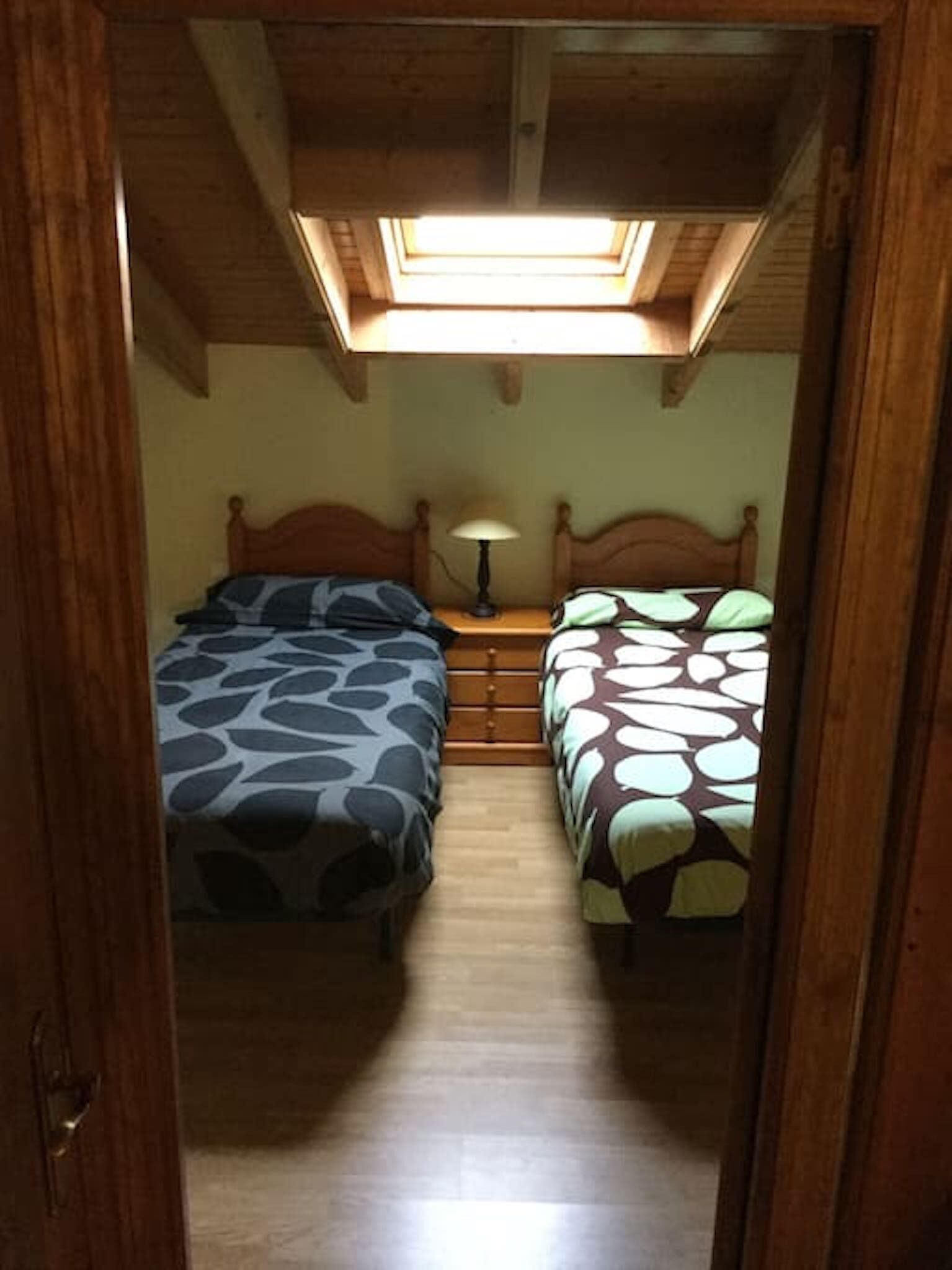 A narrow hallway leading to a bedroom with two beds, a nightstand with a lamp, and a skylight above.
