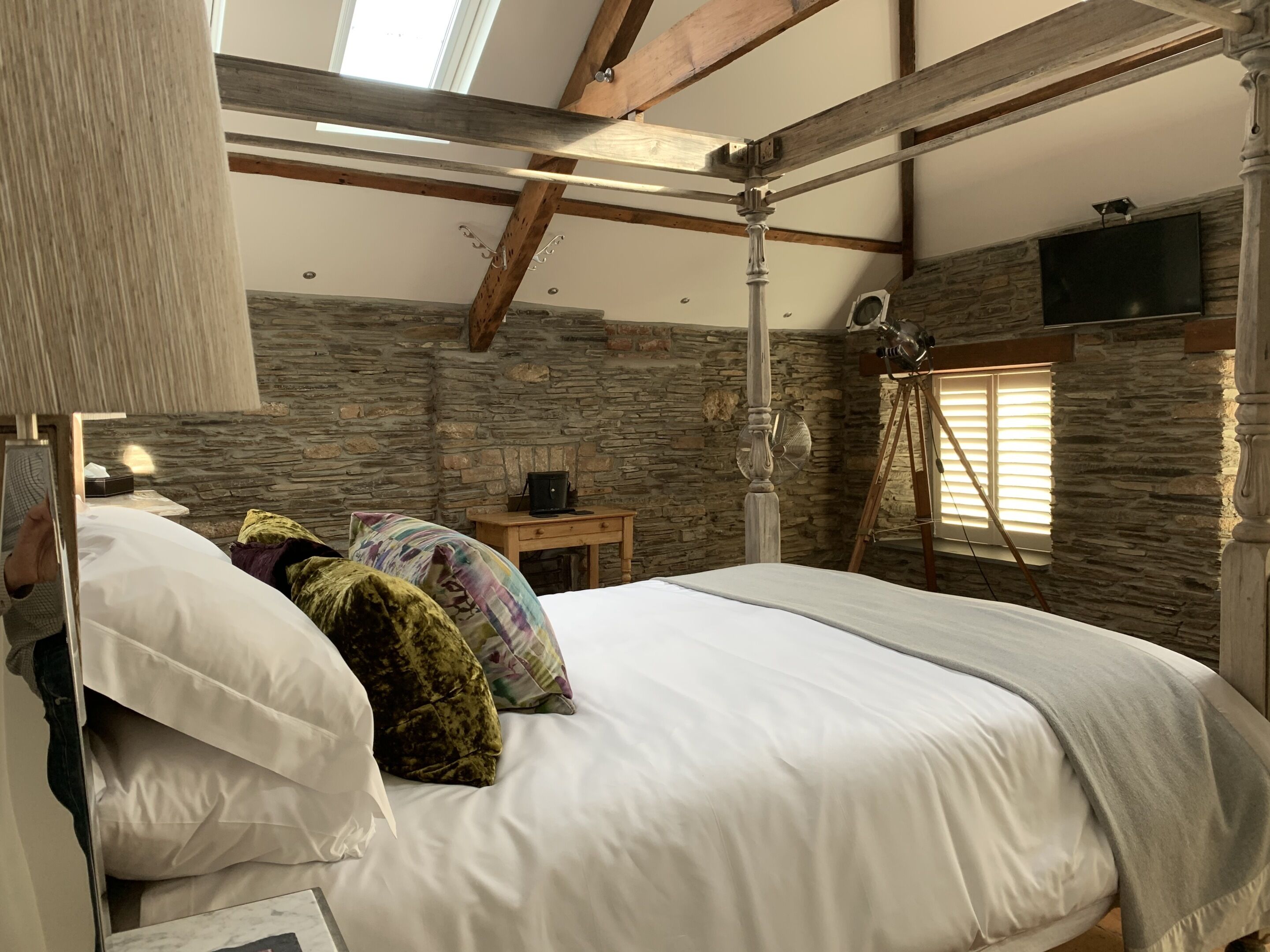 A bedroom with a stone wall, a wooden ceiling, a bed with white bedding, a wooden nightstand, and a mounted television.